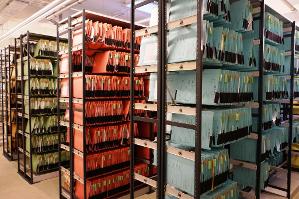 verweist auf: Verfahrensarten Aktenlager mit Regalen, in denen verschiedenfarbige Hängeakten einsortiert sind (verweist auf: Verfahrensarten)